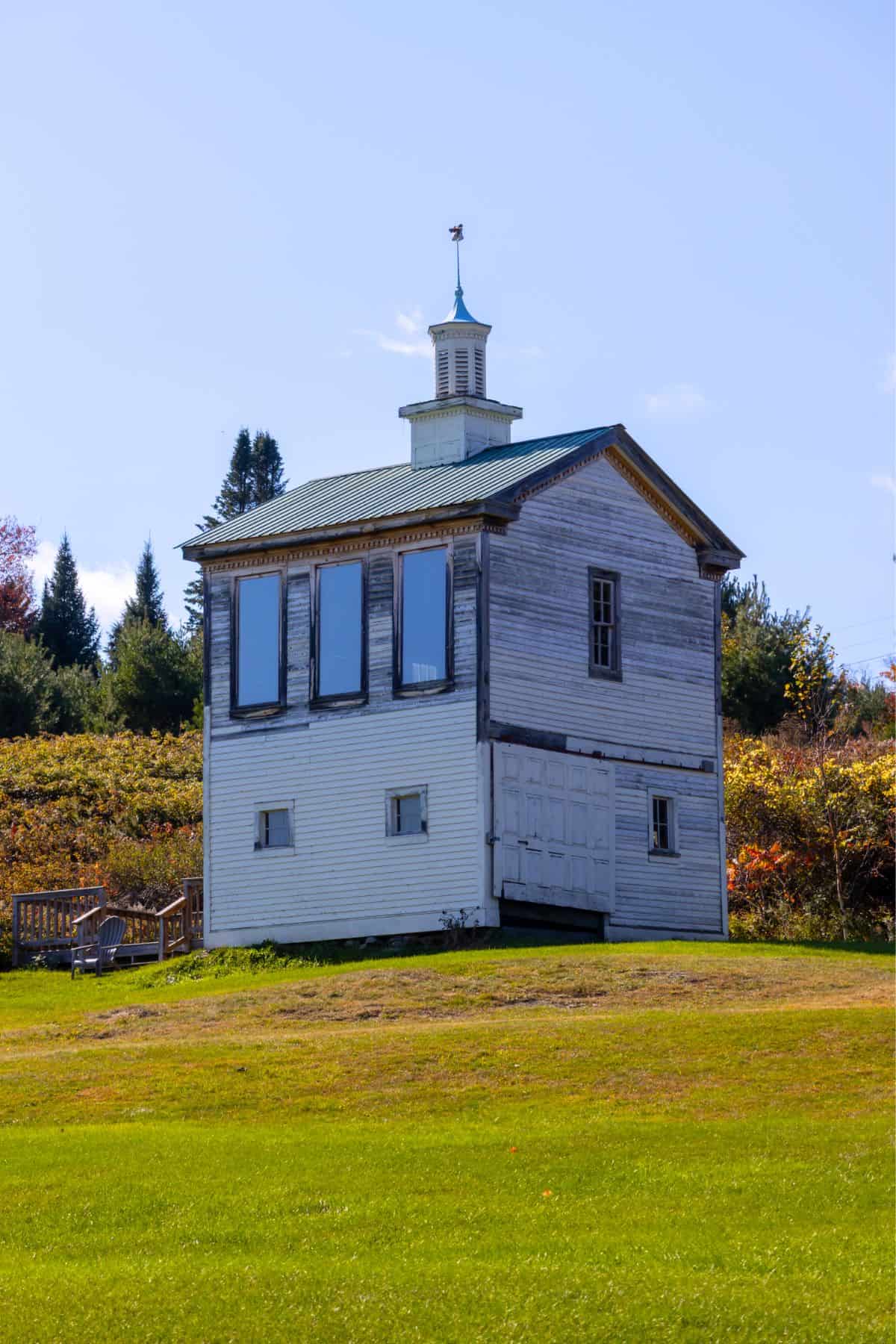 A small, weathered, white wooden building with large windows and a cupola stands on a grassy lawn with trees in the background.