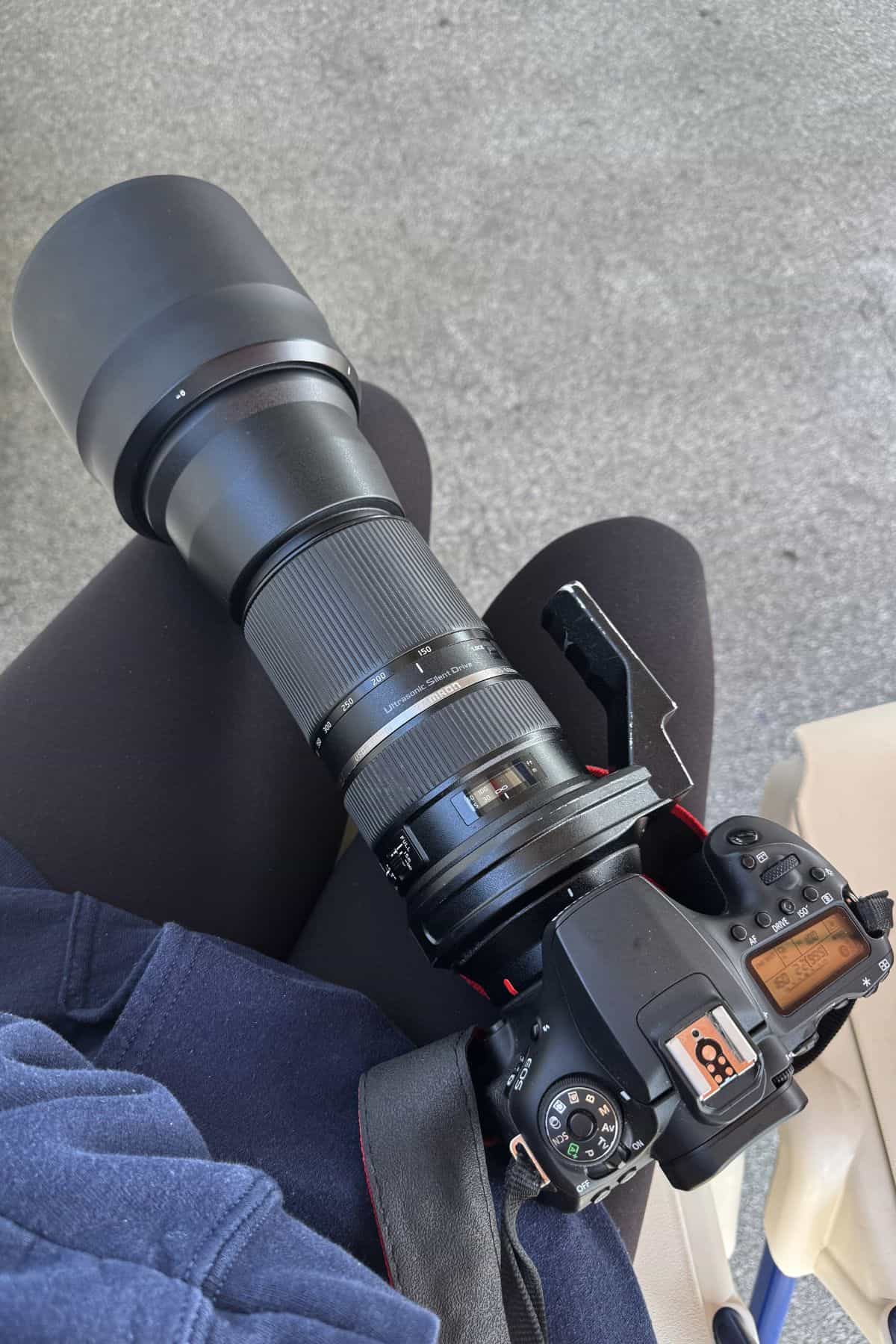 A person in dark clothing holds a DSLR camera with a long telephoto lens, resting on their lap against a gray background.