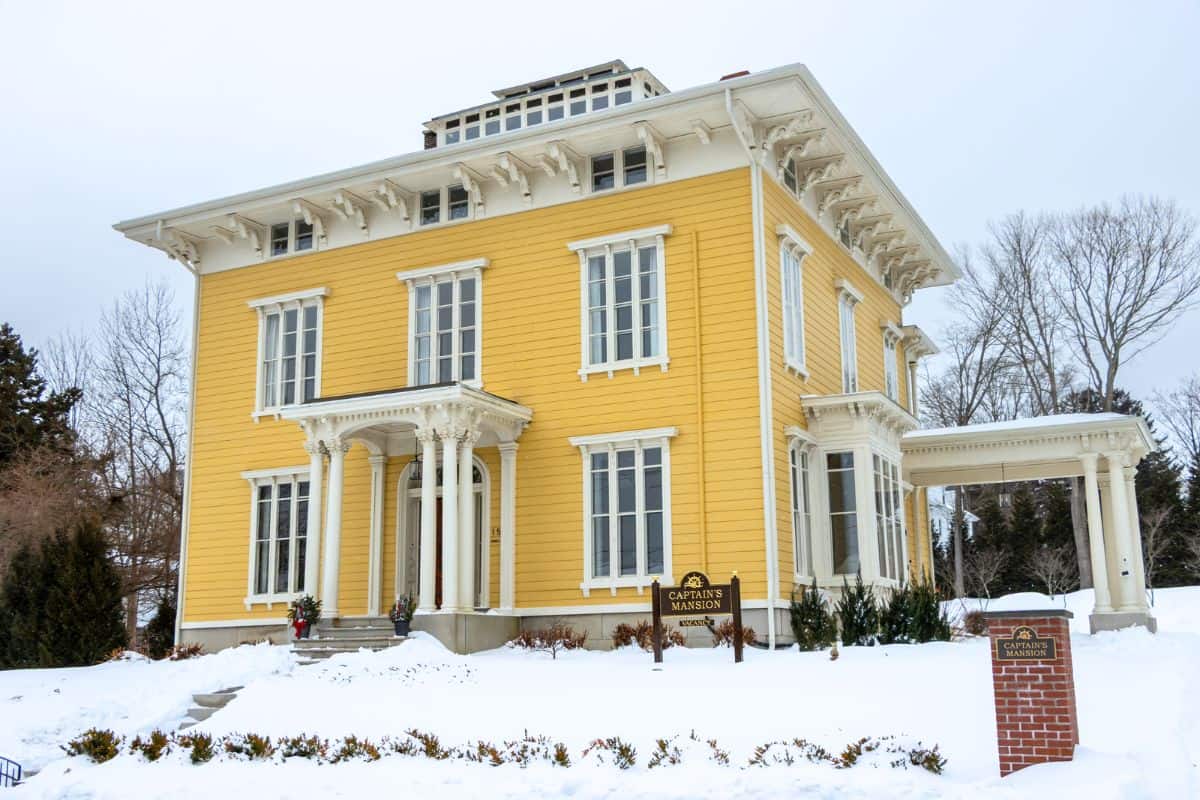 A large yellow historic house with white trim, labeled as the Caples House Museum, surrounded by snow and bare trees.