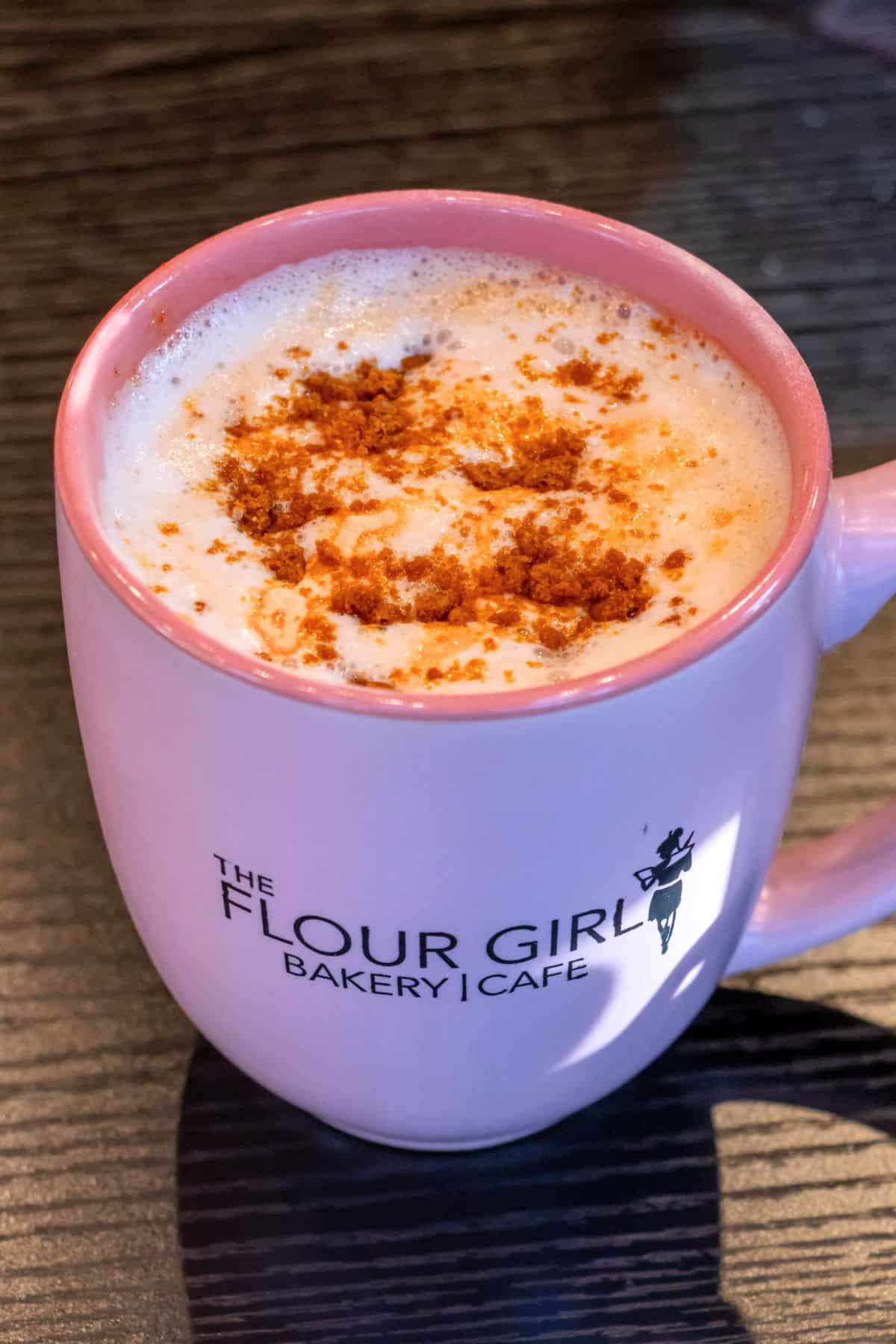 A pink mug labeled "The Flour Girl Bakery | Cafe" filled with a frothy beverage topped with crumbled brown topping sits on a dark wooden surface.