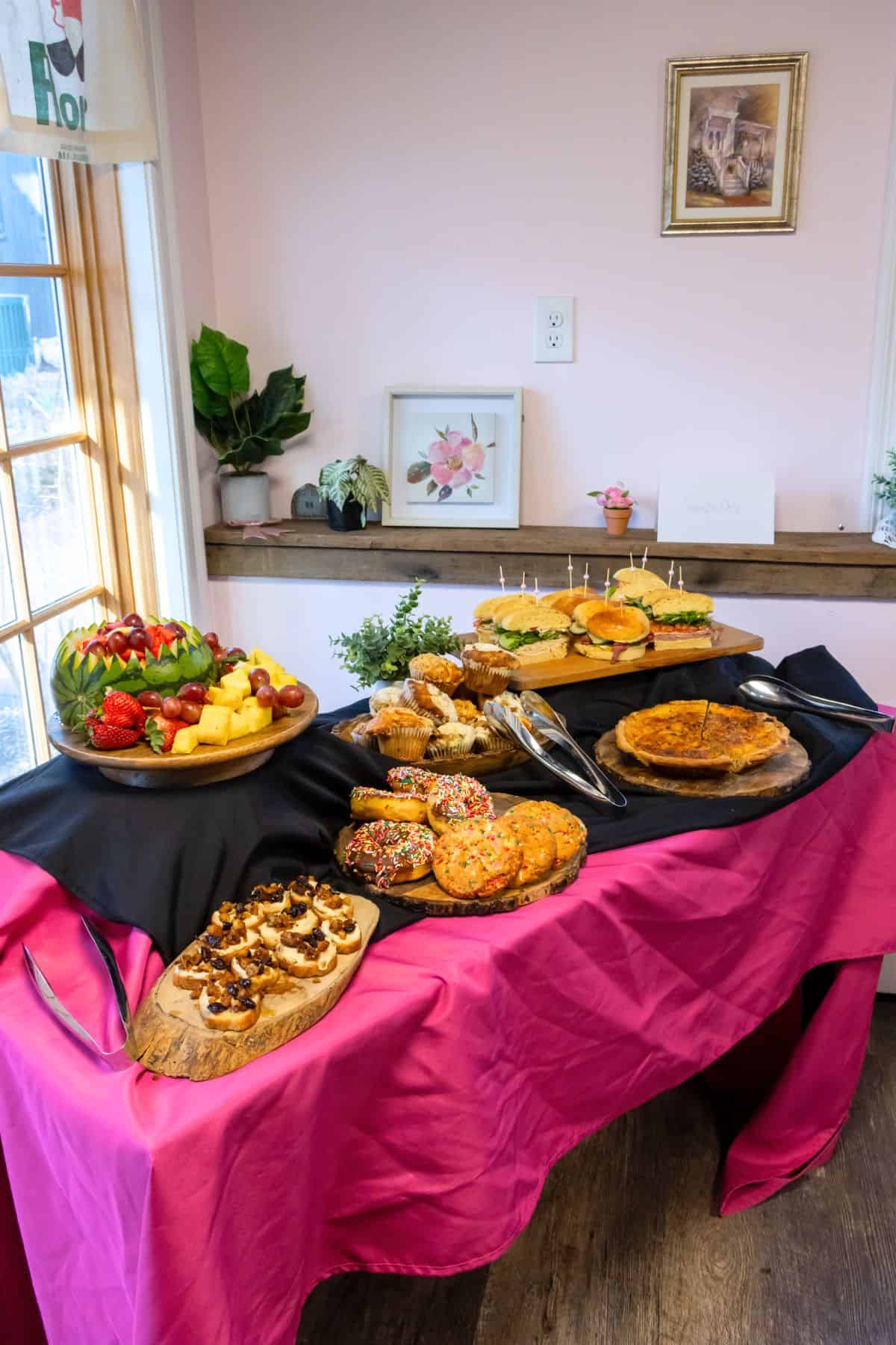 A table with a pink tablecloth displays assorted sandwiches, quiche, pastries, cookies, fruit, and appetizers, with tongs and decorative plants beside them.