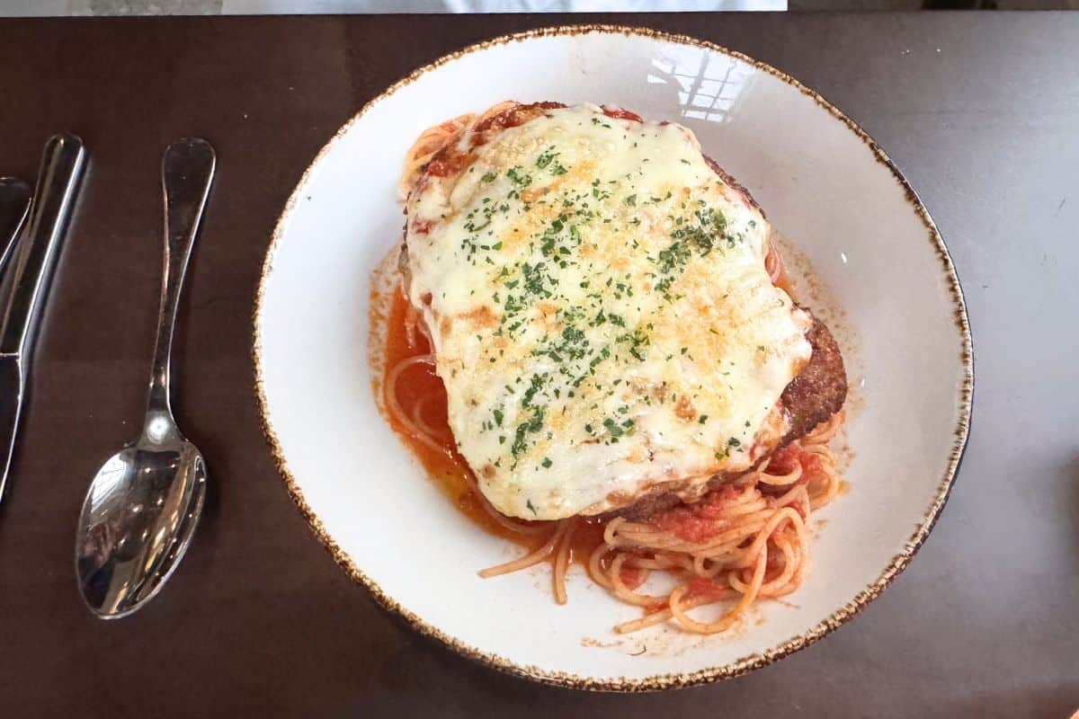 A plate of spaghetti topped with breaded chicken, marinara sauce, and melted cheese, garnished with herbs, is on a brown table next to a fork, knife, and spoon.