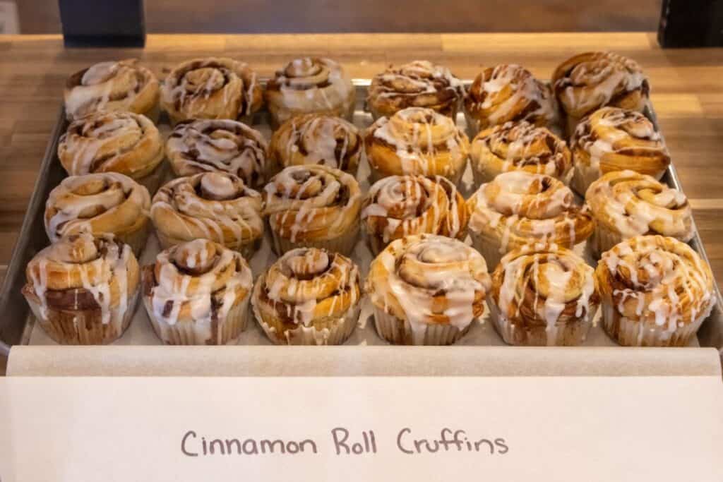 A tray of cinnamon roll cruffins with icing displayed on a wooden counter, labeled "Cinnamon Roll Cruffins.