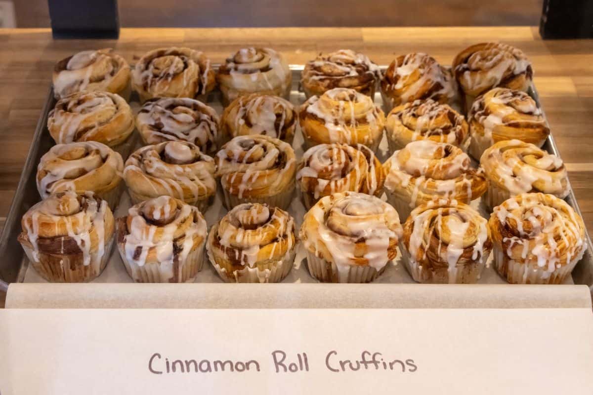 A tray of cinnamon roll cruffins with icing displayed on a wooden counter, labeled "Cinnamon Roll Cruffins.