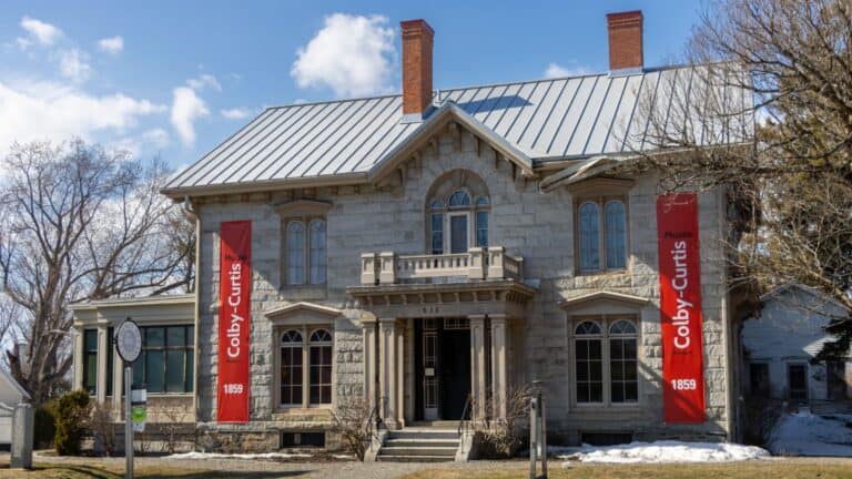 A historic stone building with two red banners reading "Colby-Curtis 1859" hanging on either side of the entrance. The building has a metal roof and a small balcony above the door.