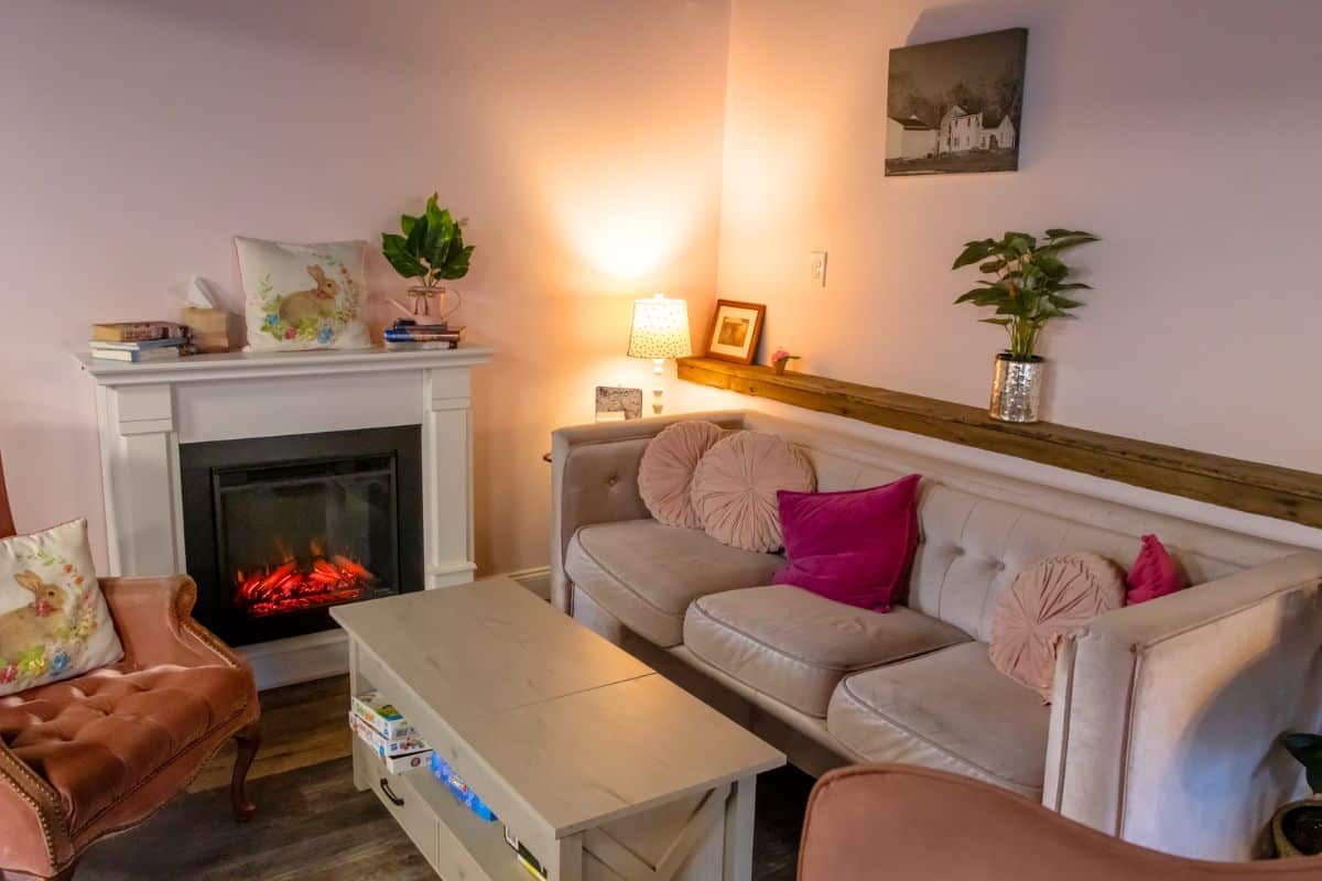 A cozy living room with a beige sofa, pink and magenta pillows, an electric fireplace, houseplants, a coffee table, and a small lamp on the mantle.