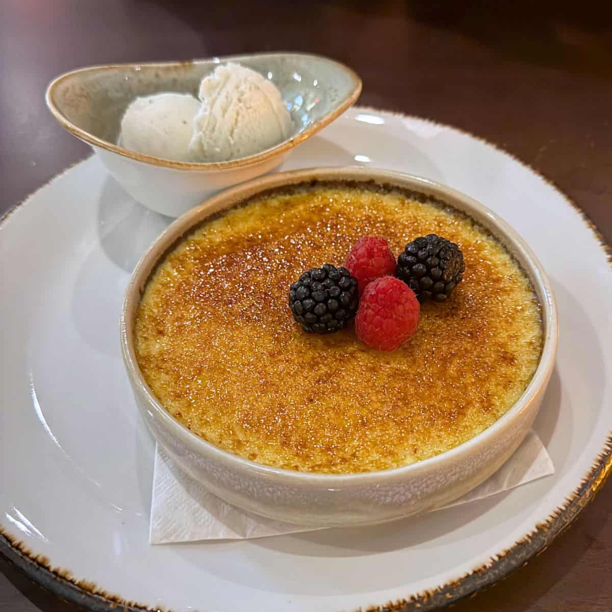 A dish of crème brûlée topped with blackberries and raspberries, served with two scoops of vanilla ice cream in a separate bowl.