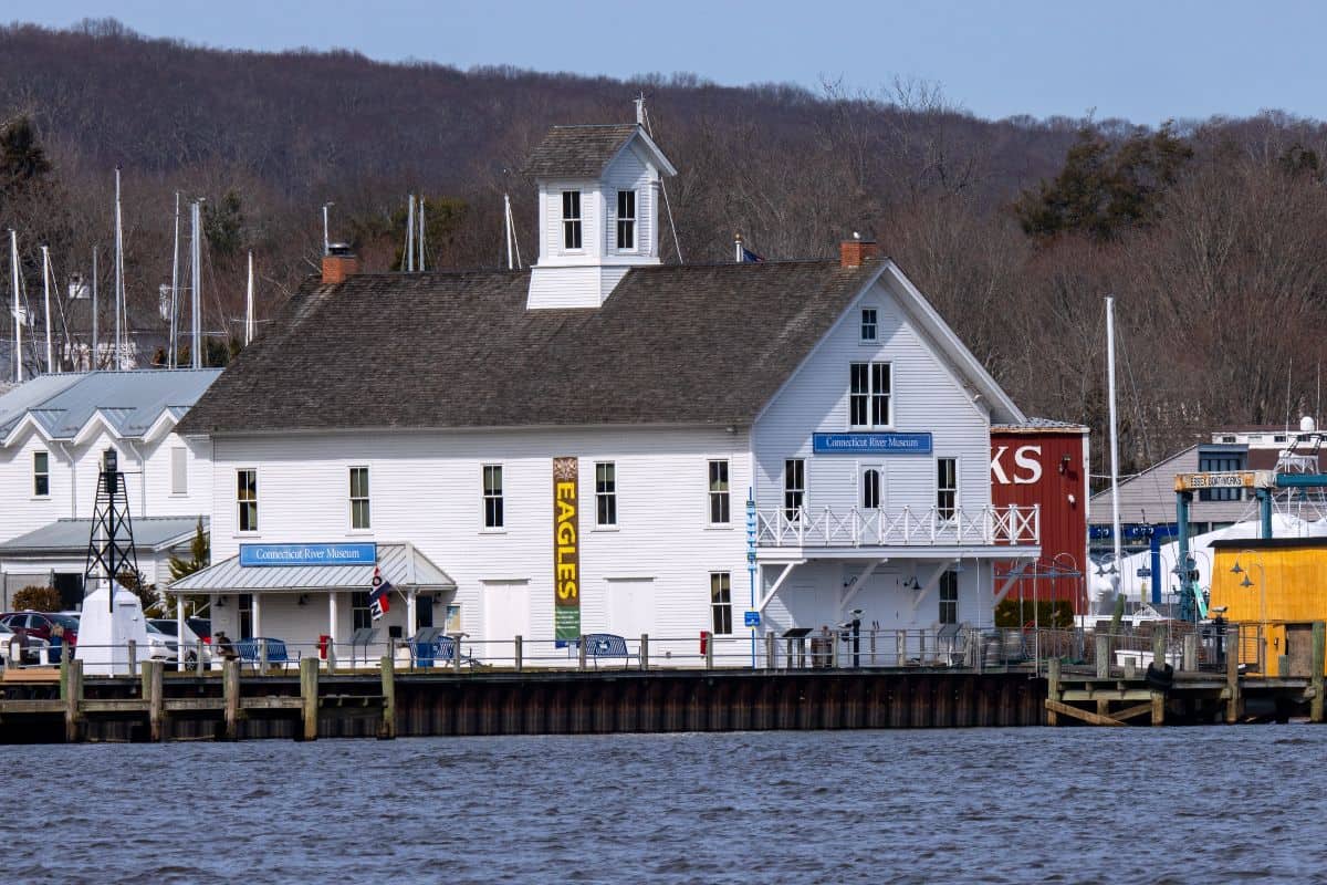 A large white waterfront building with a cupola, "EAGLES" sign, and balconies sits by a marina with sailboats and wooded hills in the background.