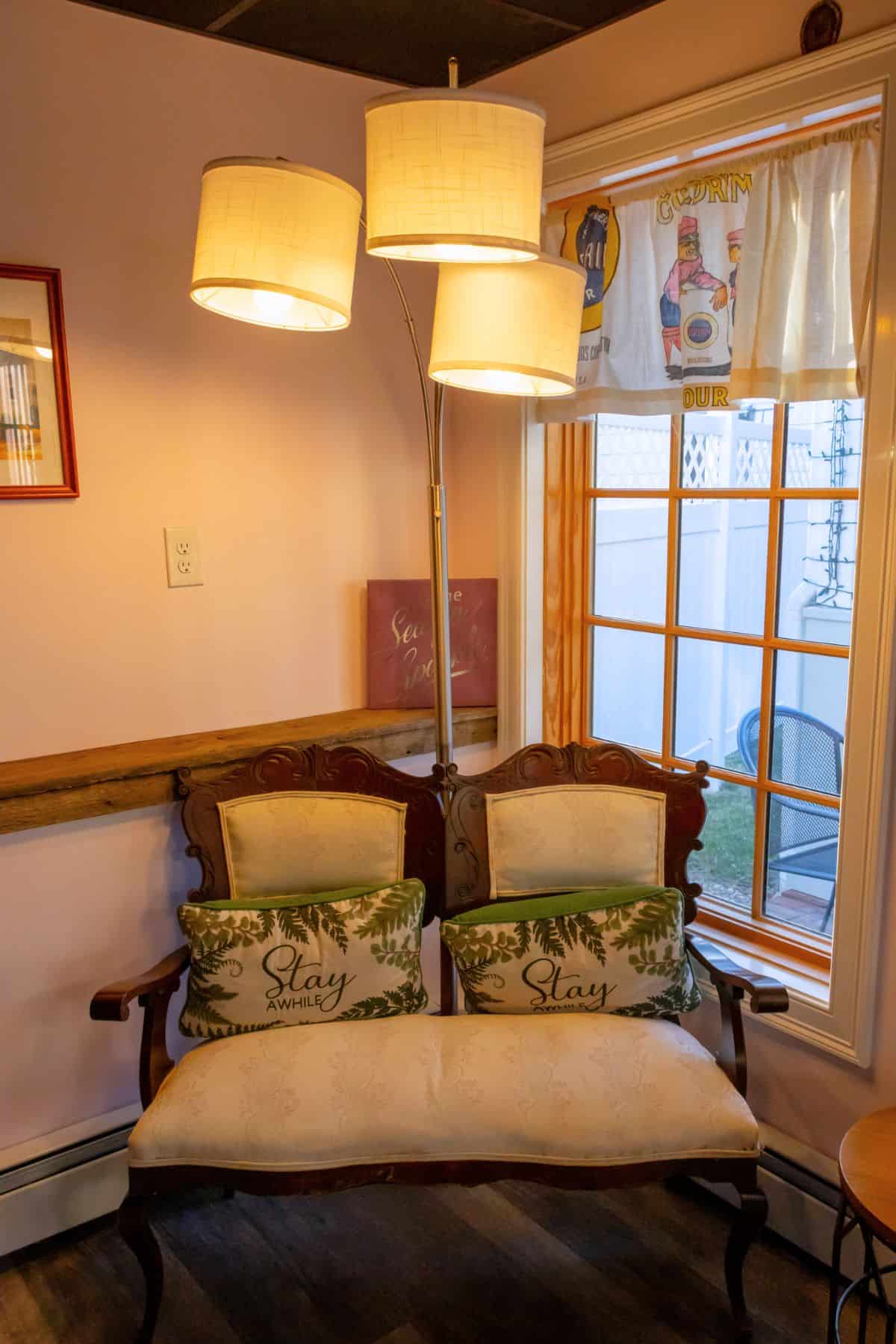A small wooden loveseat with white upholstery and two "Stay awhile" pillows sits beneath a three-shade floor lamp by a window with sheer curtains in a cozy room.