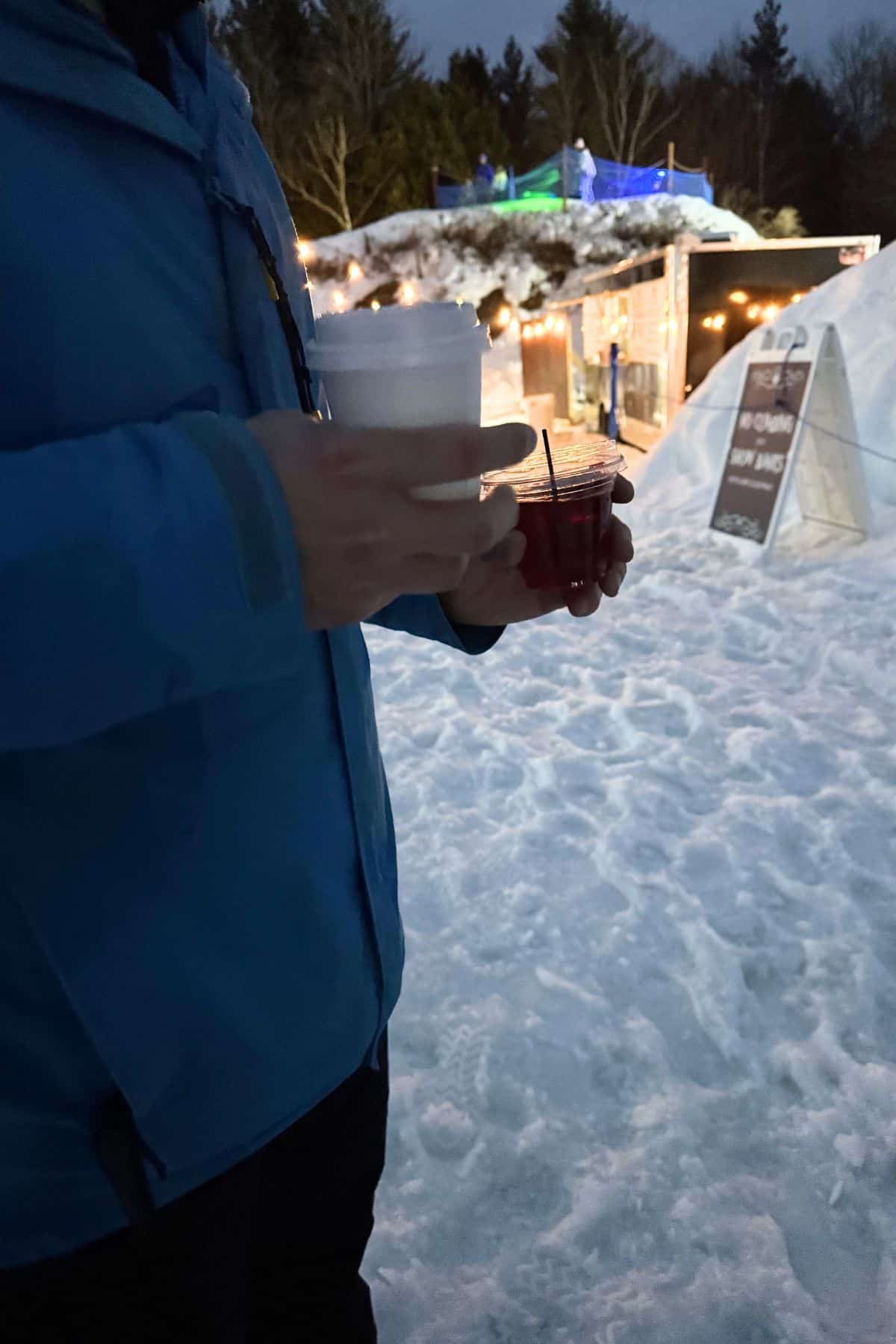 Person in a blue jacket holding two drinks, one in a white cup and one in a clear cup, while standing outdoors on snow near illuminated winter structures.