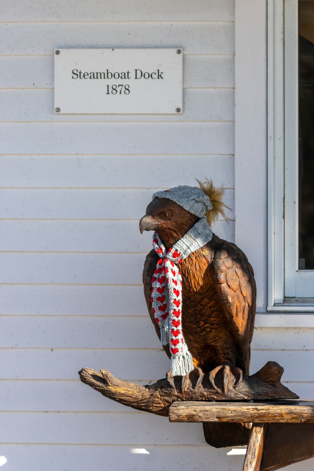 A wooden eagle statue wearing a gray knit hat and scarf sits beneath a sign reading "Steamboat Dock 1878" on a white wall.