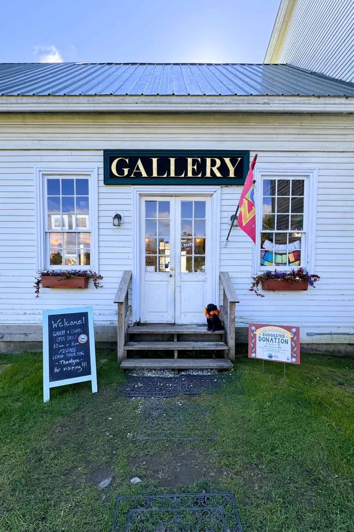 A white building with a metal roof and a sign reading "GALLERY" above double doors, flanked by a chalkboard welcome sign and a donation sign. Colorful flags are displayed by the entrance.