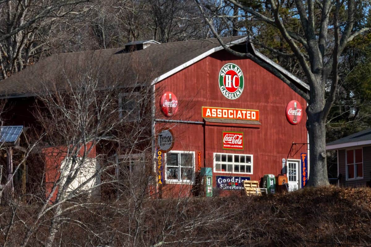 A red wooden building with vintage signs for Coca-Cola, Goodyear, and Associated Gasoline, partially obscured by leafless trees.