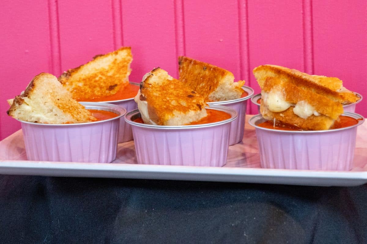 Five ramekins filled with tomato soup, each topped with a grilled cheese sandwich wedge, are arranged on a tray in front of a pink wall.