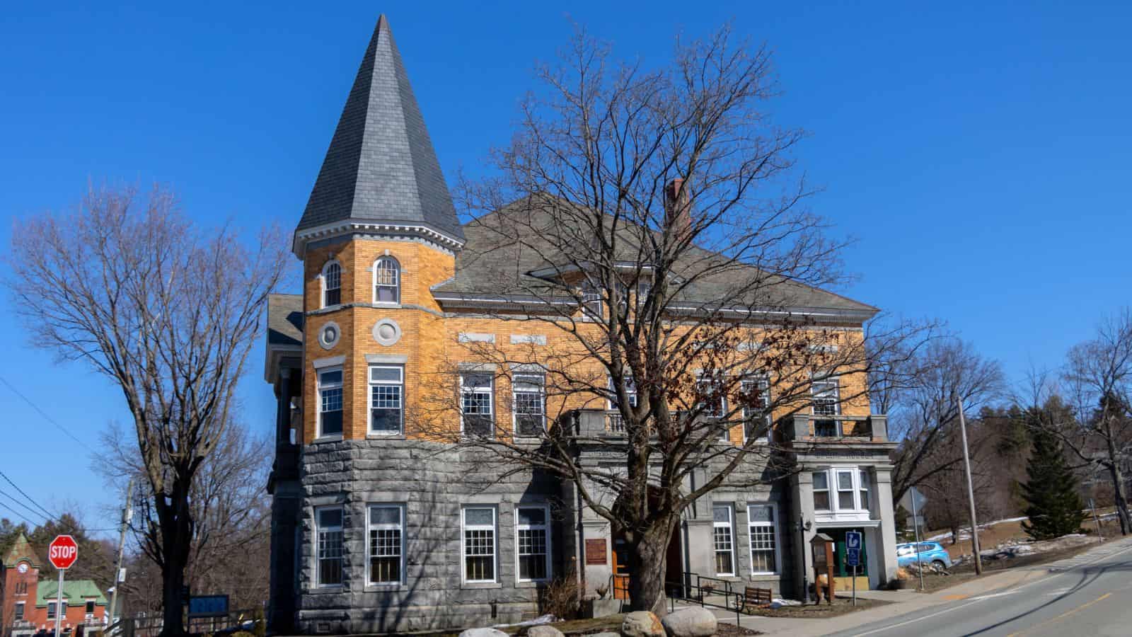 A historic brick and stone building with a tall, pointed tower and leafless trees in front, set on a clear, sunny day.