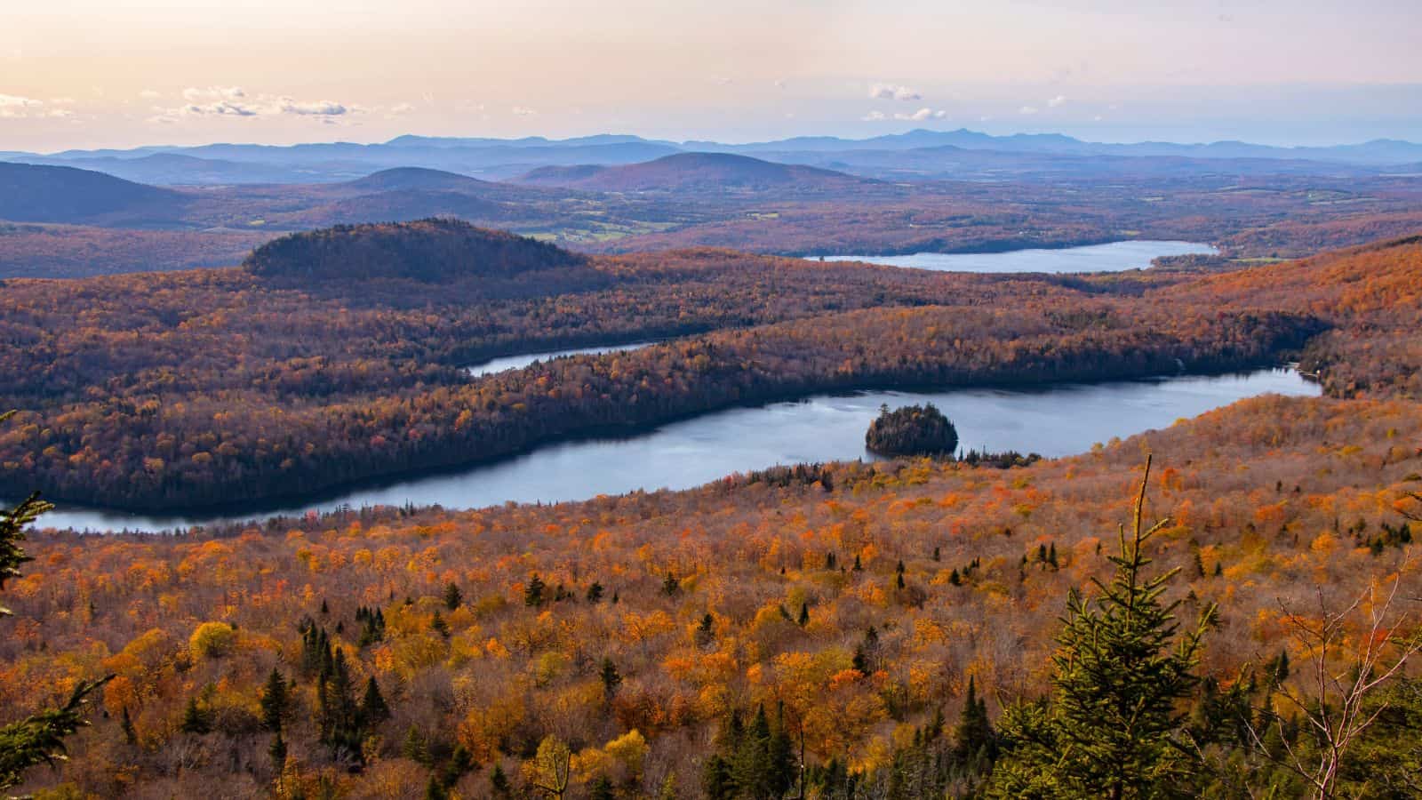 A panoramic view of a forested landscape in autumn with lakes, rolling hills, and distant mountains under a clear sky.