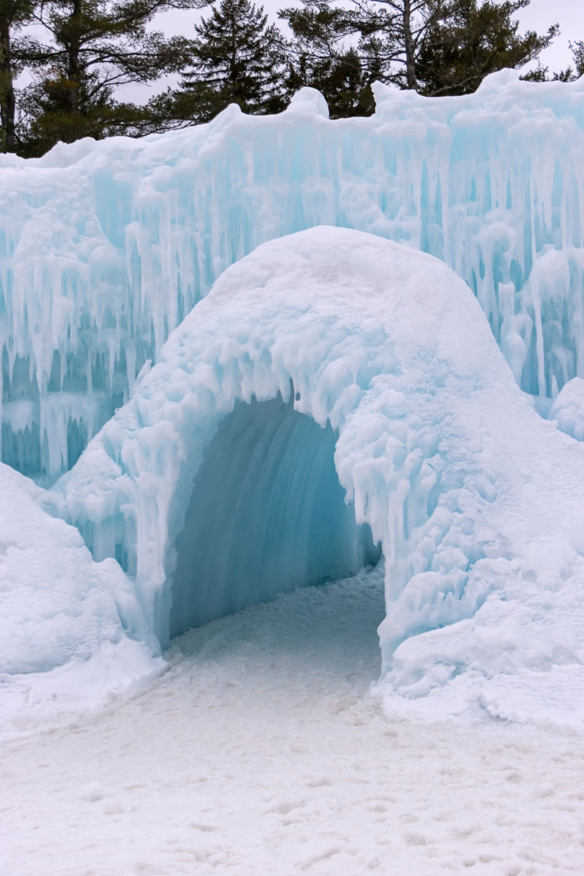An entrance tunnel made of snow and ice, with icicles hanging above, leads into a larger ice formation; trees are visible in the background.