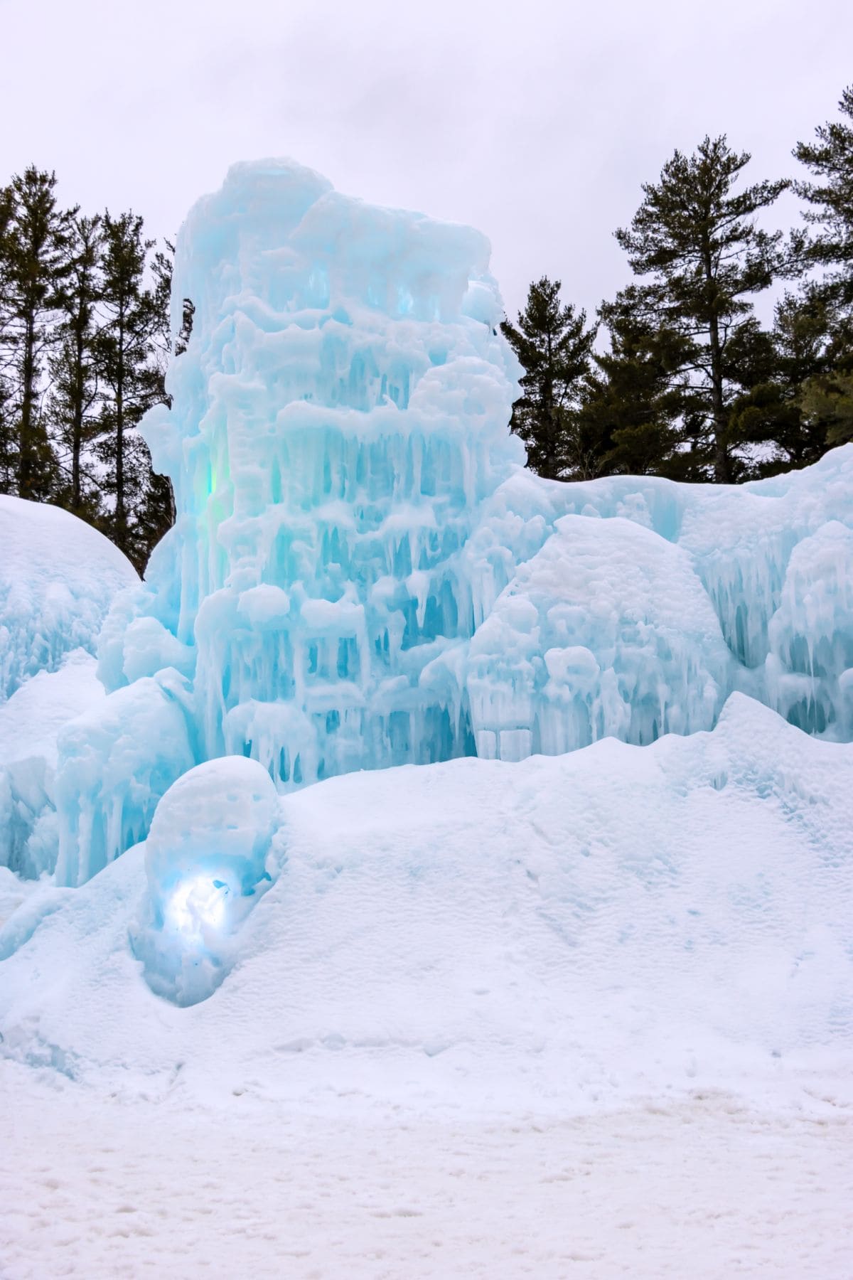 A tall, jagged formation of blue-tinted ice stands amid snow with pine trees in the background under a cloudy sky.