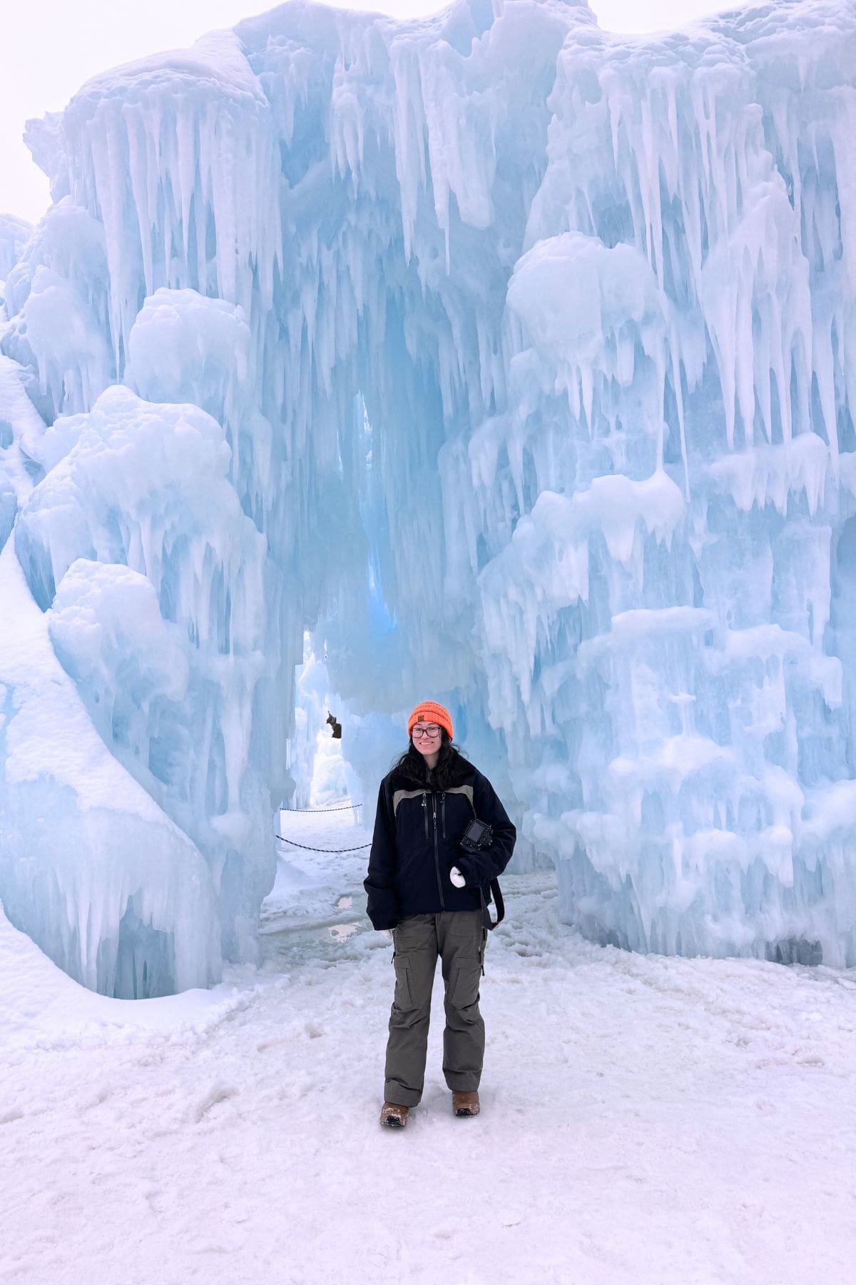 A person in winter clothing stands in front of a large ice formation with icicles and an archway, surrounded by snow.