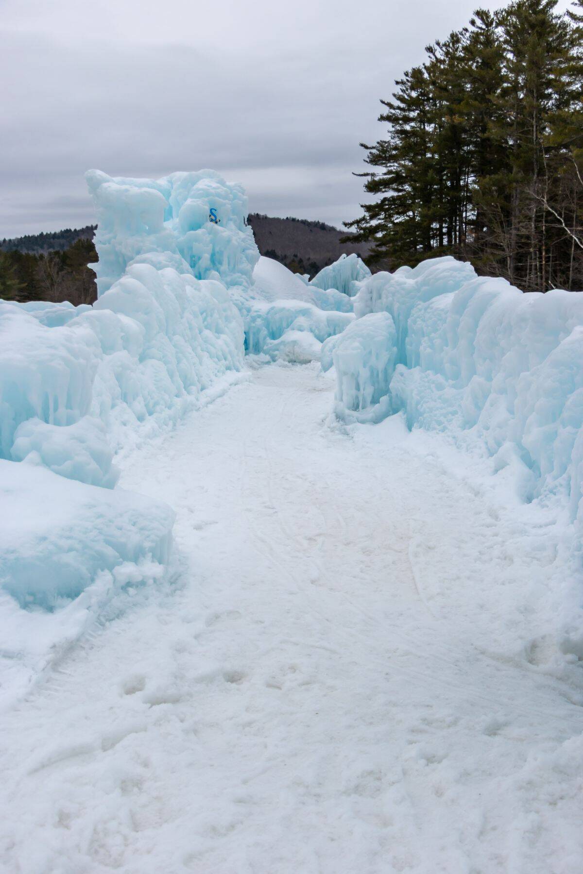 A snow-covered path passes between tall walls of ice, with evergreen trees and hills visible in the background under a cloudy sky.