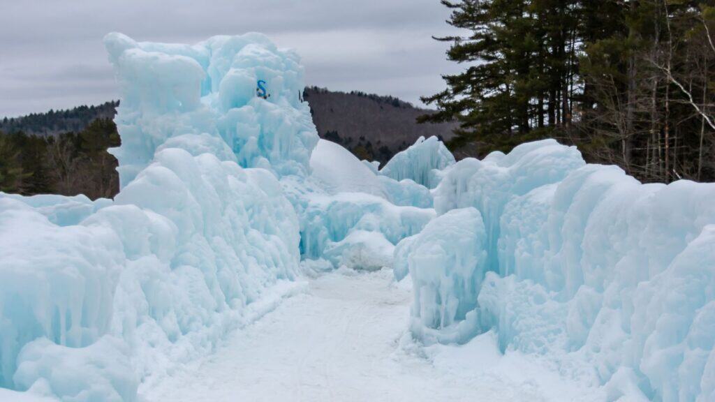 A winter path lined with thick, sculpted blue ice formations, surrounded by trees and distant hills under a cloudy sky.