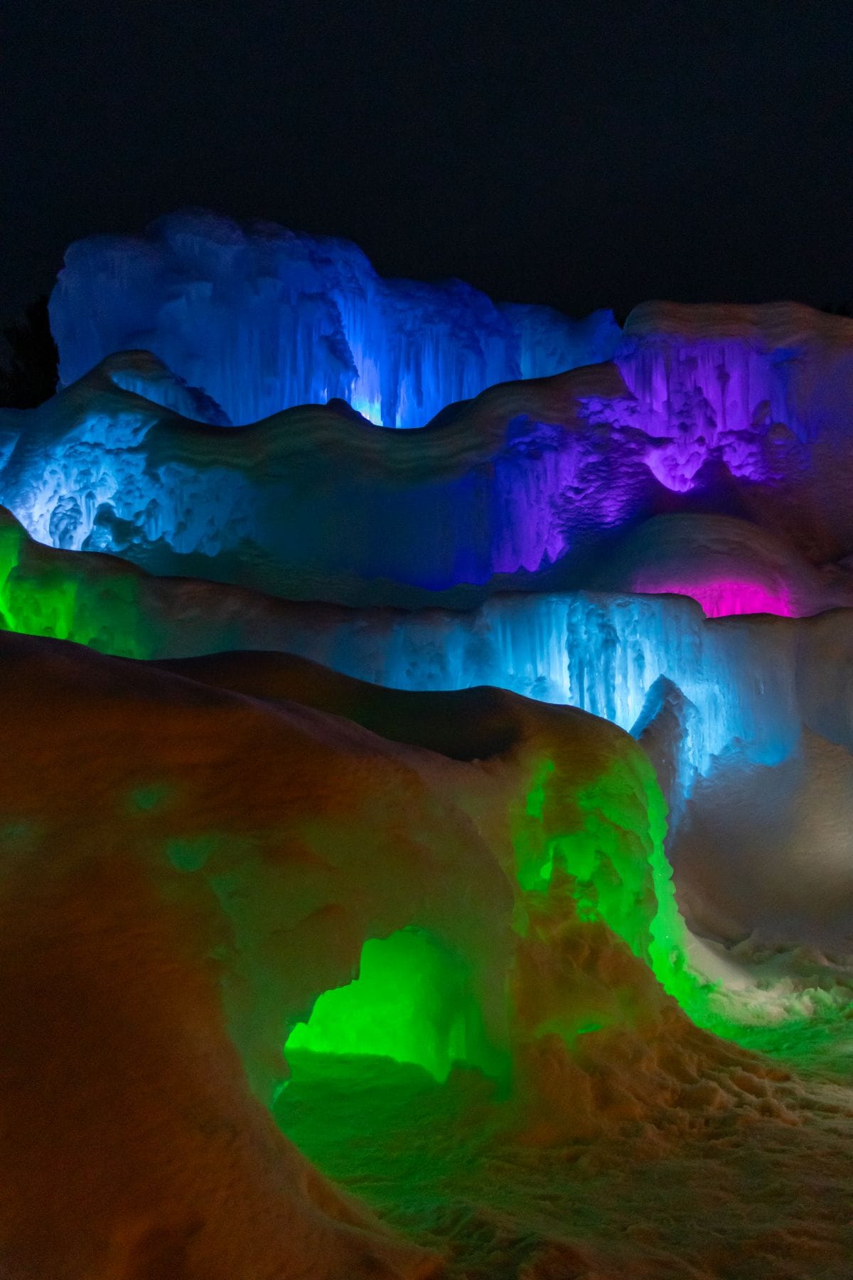 Multicolored lights illuminate large, layered ice formations at night, displaying shades of blue, purple, and green against a dark sky.