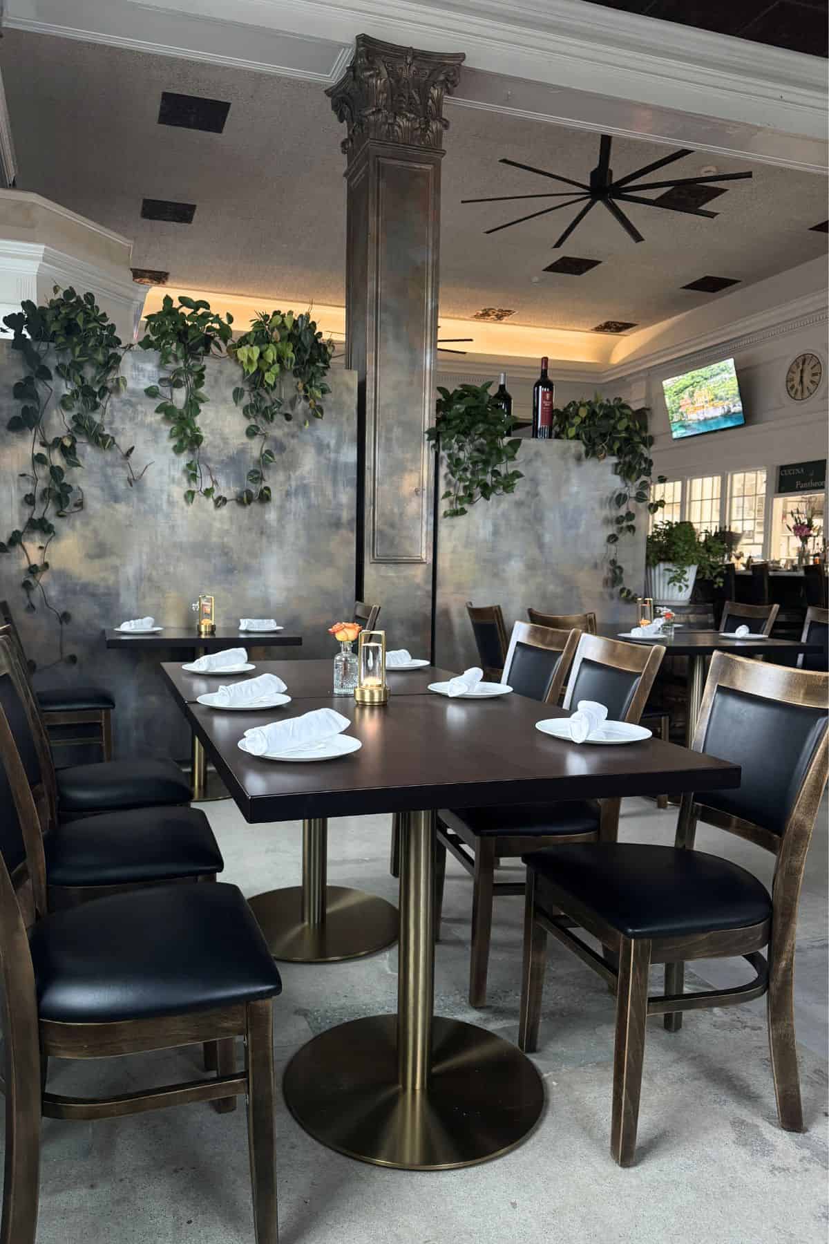A restaurant dining area with dark wooden tables and chairs, neatly rolled white napkins, leafy plants, and a large pillar in the center. A TV is mounted on the wall in the background.