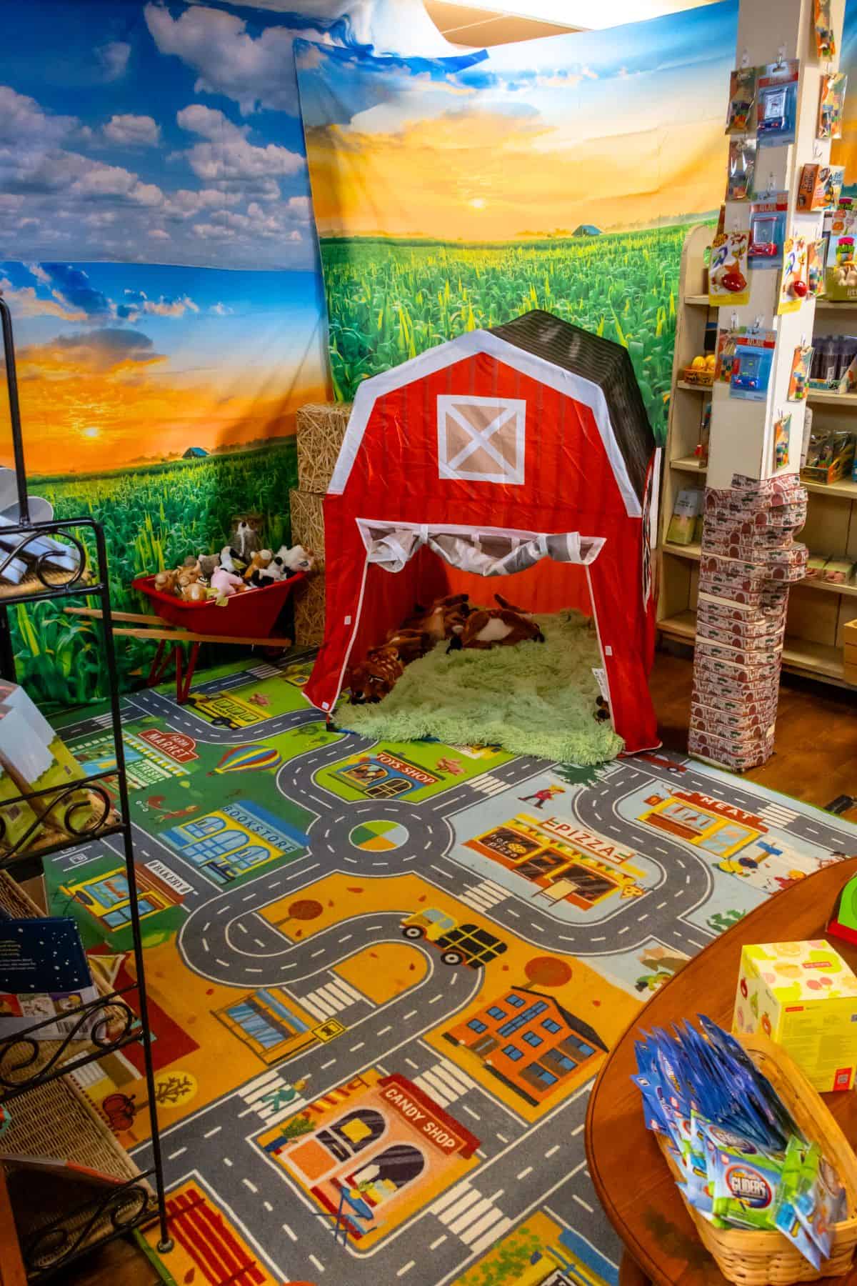 Children’s play area with a red barn tent, hay, toy animals, colorful play mat with roads, and shelves of toys in a room with a farm-themed mural.