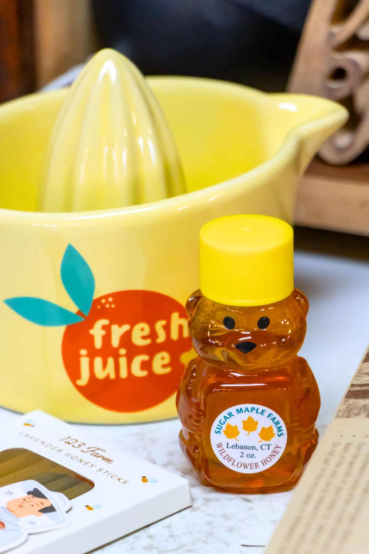 A small bear-shaped honey bottle labeled "Sugar Maple Farms" sits in front of a yellow citrus juicer with "fresh juice" written on it.