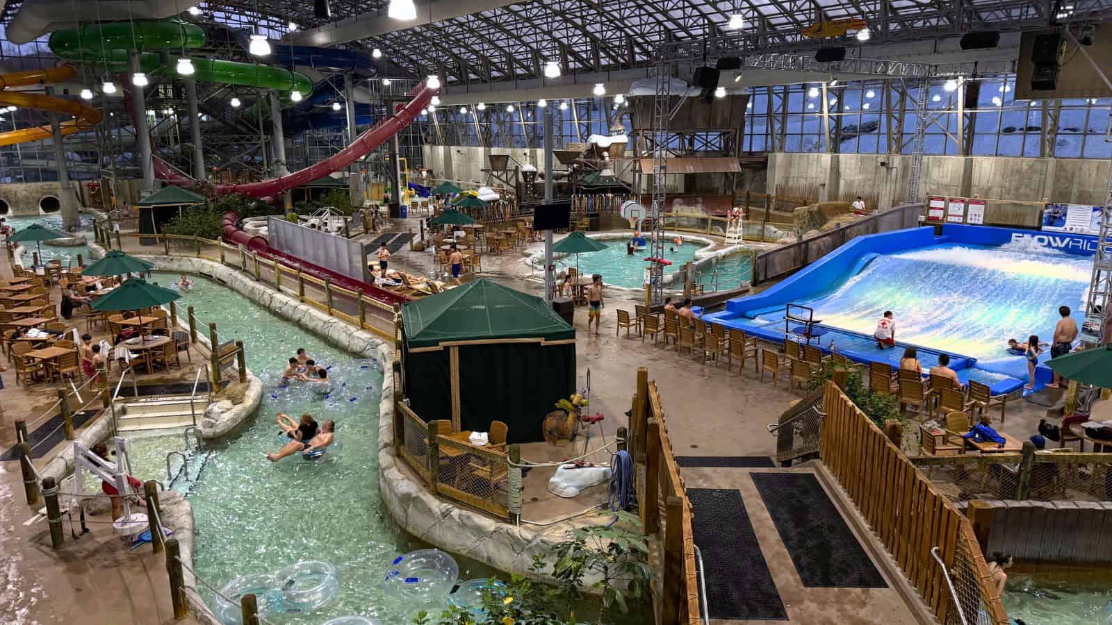 Indoor water park with a lazy river, wave simulator, swimming pools, water slides, lounge chairs, and people enjoying various attractions under a large glass roof.
