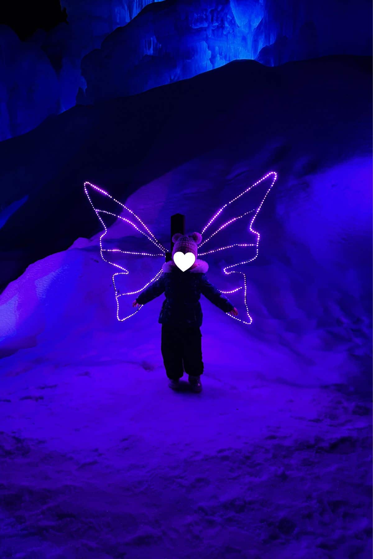 Person standing in the snow at night, illuminated by purple LED lights shaped like butterfly wings, with a heart sticker over their face. Blue-lit ice structures are visible in the background.