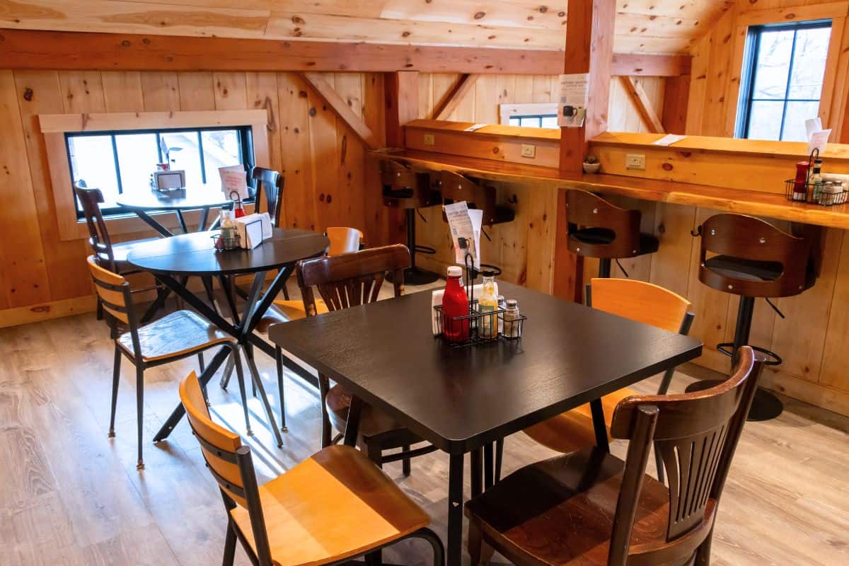 Cozy cafe interior with wooden walls, several tables and chairs, a counter with stools by the window, and condiments on the tables.