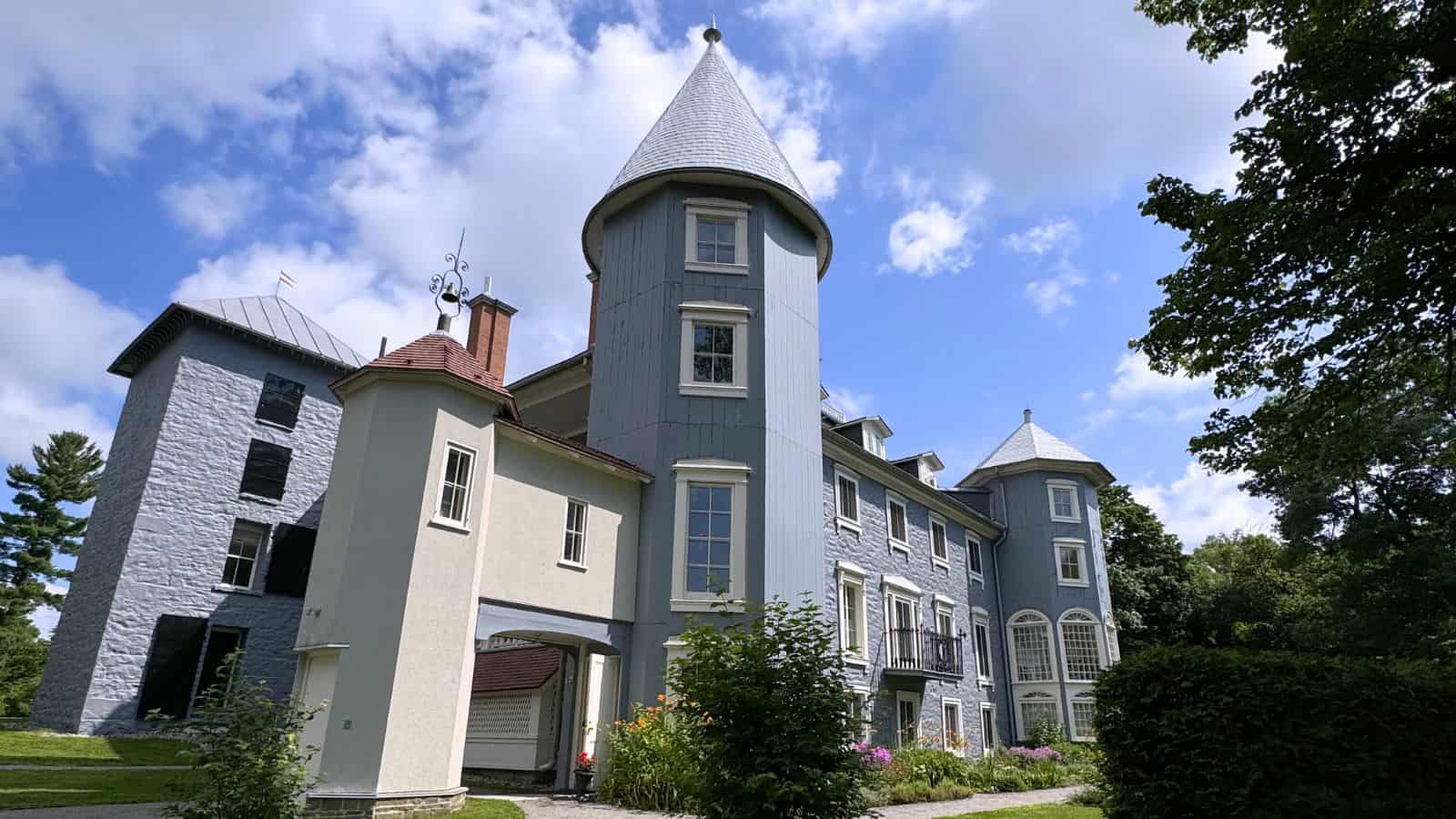 Large, light blue and white building with turret, surrounded by greenery and garden, under a partly cloudy sky.