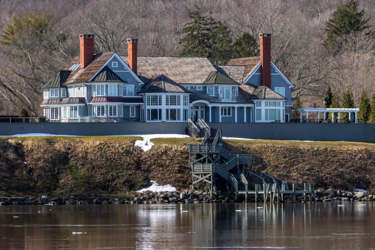 Large waterfront house with multiple chimneys, surrounded by bare trees, a wooden staircase leading to a dock, and patches of snow on the ground.