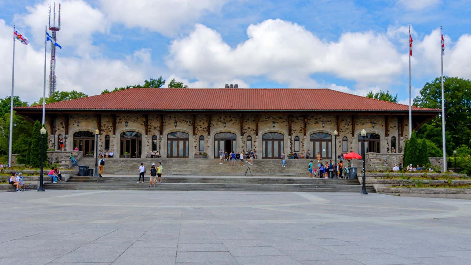 A large stone building with a red tile roof, wide steps, and people sitting or standing outside on a sunny day. There are flags and trees around the building.