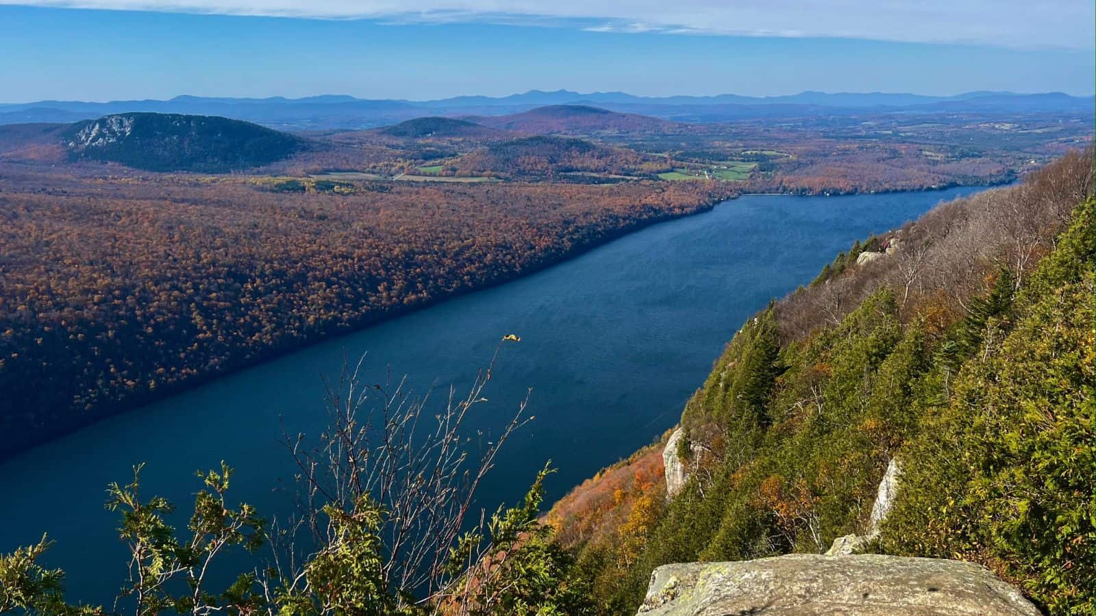 A wide river winds through a forested valley in autumn, with hills and distant mountains under a clear blue sky.