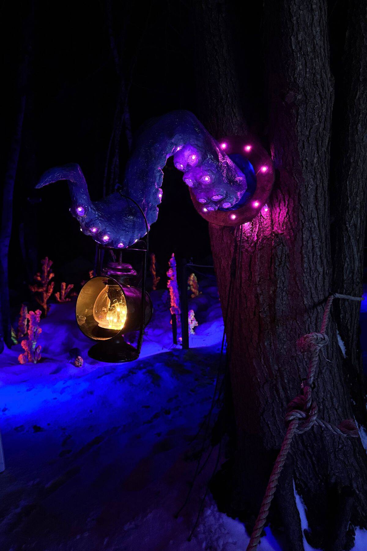 A tree trunk with a large, purple-lit tentacle sculpture wrapped around it holds a lantern; snow and illuminated decorations are visible in the background.