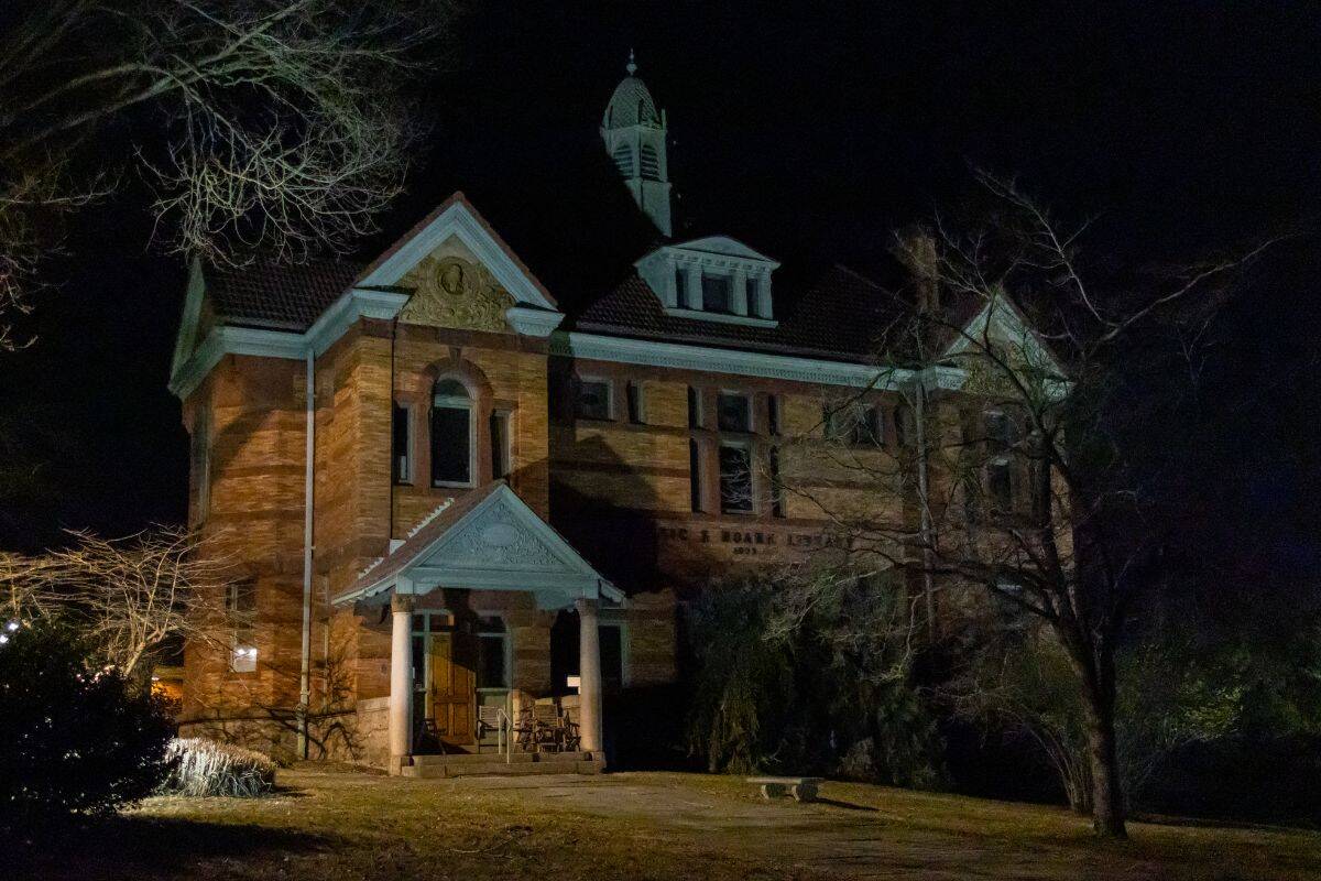 A large brick building with a porch and steeple is illuminated at night, surrounded by bare trees and dark shadows.