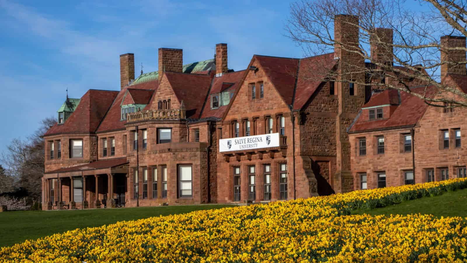 A large red-brick building with a sign reading "Salve Regina University," surrounded by a field of blooming yellow flowers under a clear blue sky.