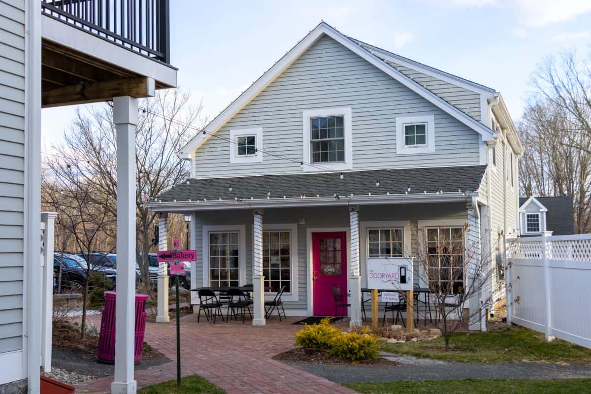 A small, gray two-story building with white trim houses a cafe; outdoor tables and chairs line the brick walkway leading to the entrance.