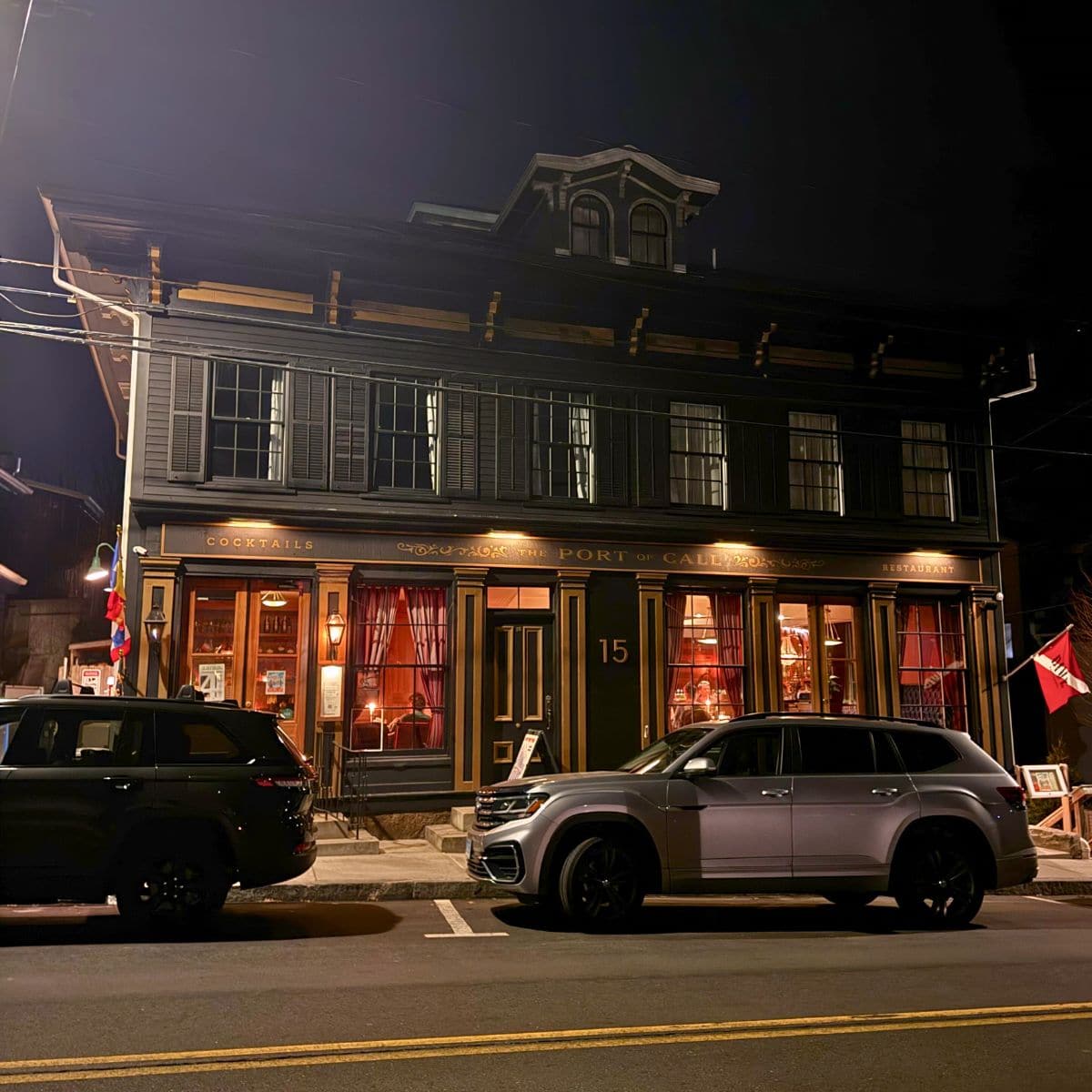Two cars are parked on the street in front of a dark, historic building with large windows illuminated from inside at night.