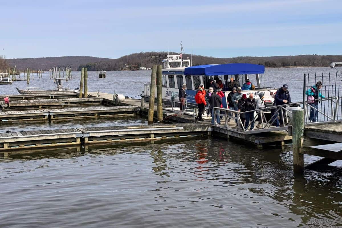 A group of people disembark from a small passenger boat at a dock on a calm river, with hills and empty docks visible in the background.