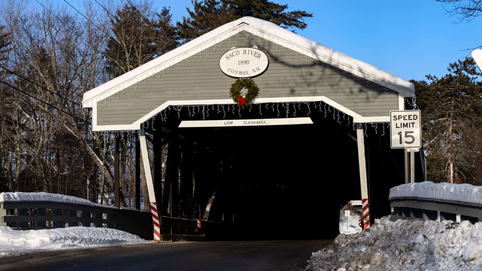 A gray covered bridge labeled "Saco River 1890, Conway, NH" with a wreath above the entrance, snow on the ground, and a speed limit sign of 15 mph nearby.