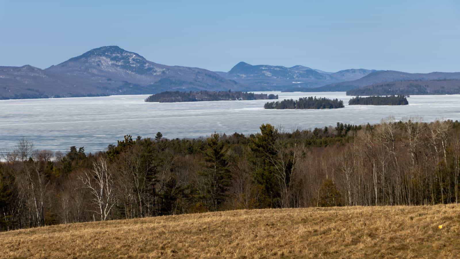 A frozen lake with several tree-covered islands, bordered by forest and rolling hills, with mountains in the background under a clear sky.