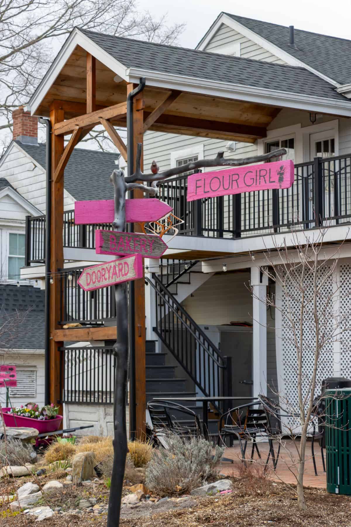 Outdoor scene with a two-story building, black staircase, and several pink directional signs reading "FLOUR GIRL," "BAKERY," and "DOORYARD" on a metal post in front.