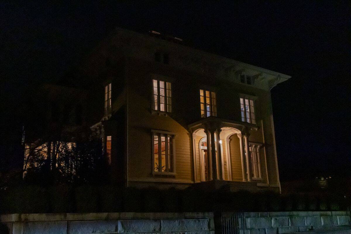 A large house with tall windows is illuminated from within at night, with a lit entrance and dark surroundings.