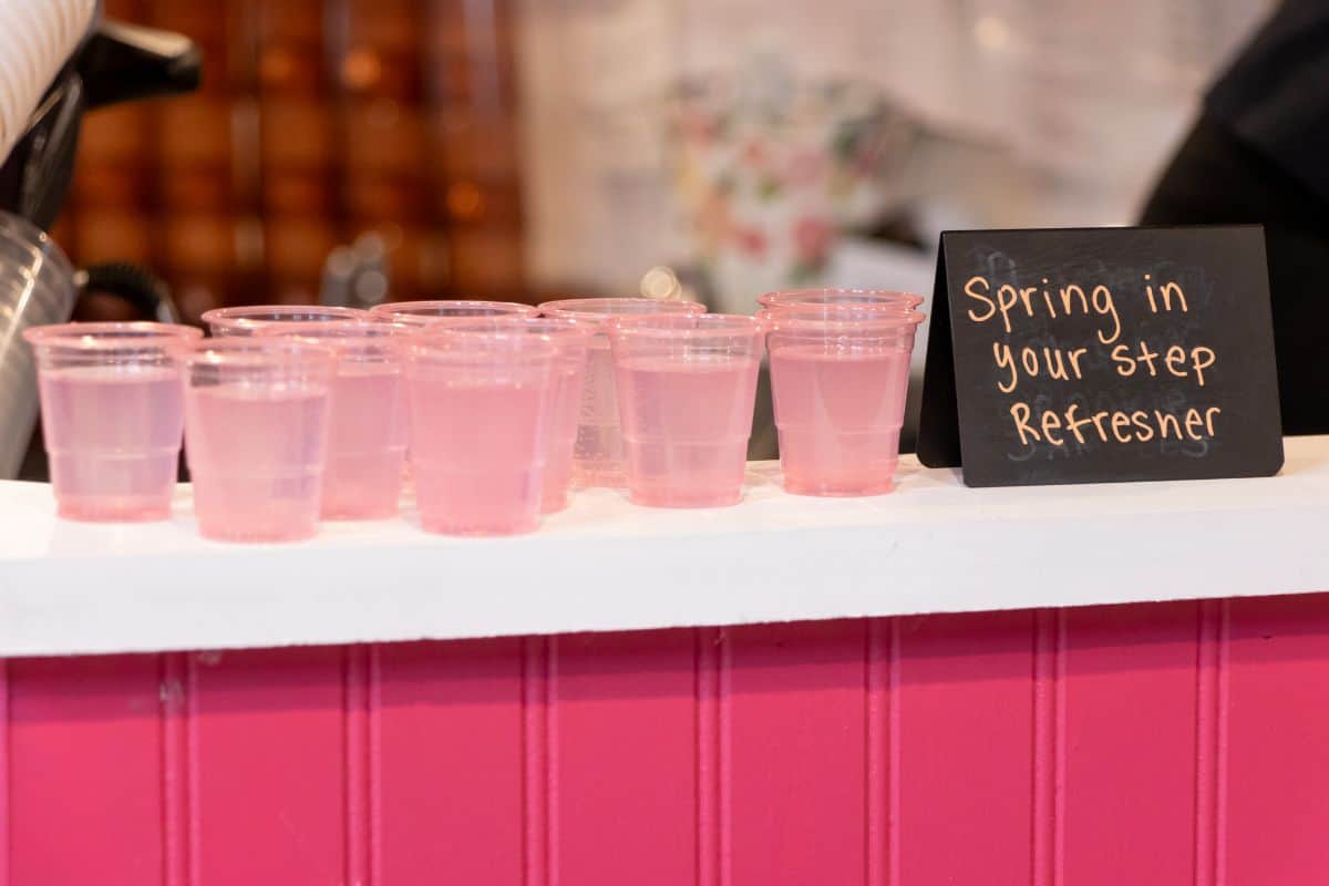 Several small plastic cups filled with pink liquid are arranged on a counter next to a chalkboard sign reading, "Spring in your step refresher.