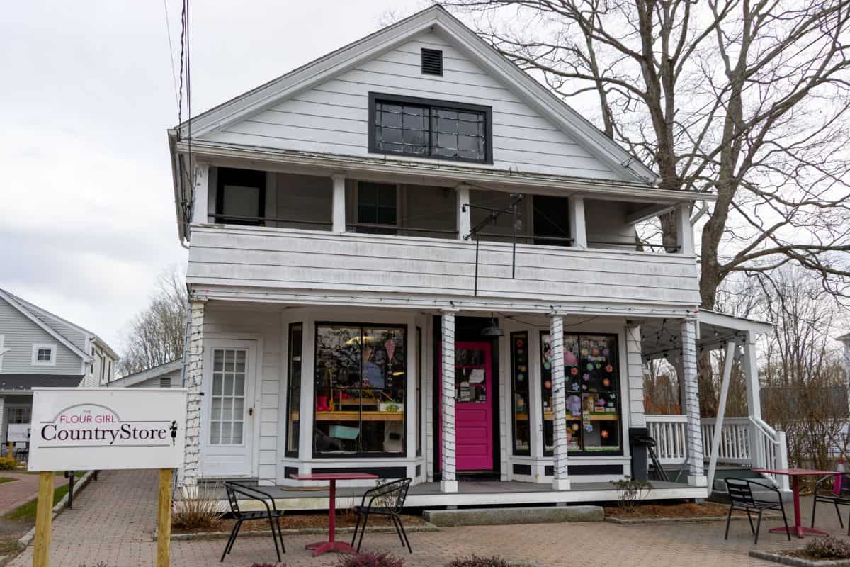 A two-story white building houses the Flour Girl Country Store, with a pink door, porch seating, and a sign in front. The trees nearby are bare, suggesting early spring or late fall.