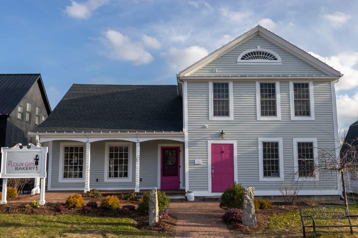 A white, two-story building with bright pink doors and shutters houses the Flour Girl Bakery, with a sign on the front lawn and clear sky overhead.