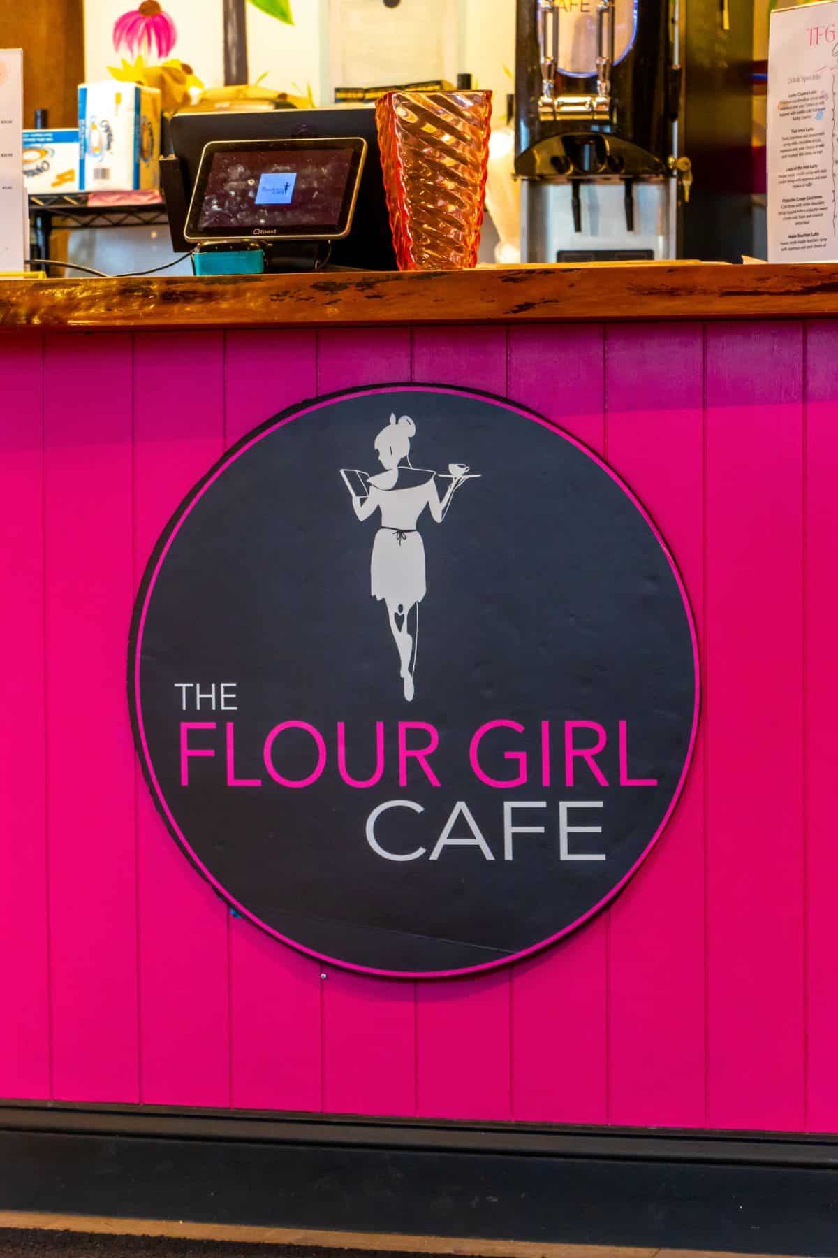 A sign reading "The Flour Girl Cafe" is displayed on a bright pink counter; the logo features a woman holding a tray.